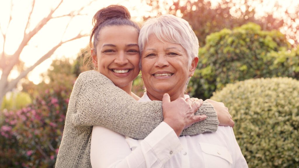 A young woman lovingly hugs an older woman from behind as they both smile warmly in a sunlit garden.